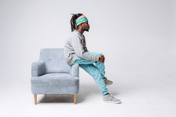 Portrait of a smiling black young model in a gray sweater and blue pants on a gray background near the chair