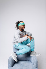 Portrait of a smiling black young model in a gray sweater and blue pants on a gray background near the chair