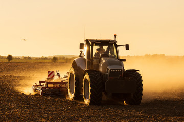 Modern tractor plowing at the sunset with lot of dust in background and with a plane taking off