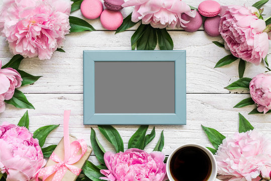 blank photo card in frame made of pink peony flowers, macarons and coffee cup on white wooden background. flat lay