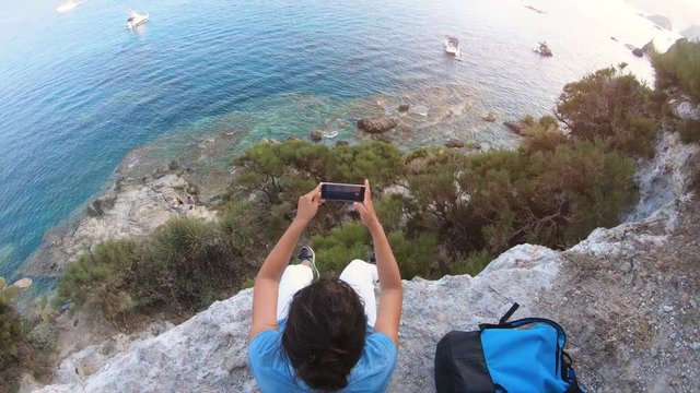 Young woman taking pictures with smartphone sitting on cliff edge looking at the sea from a bay on Ponza Island coast.