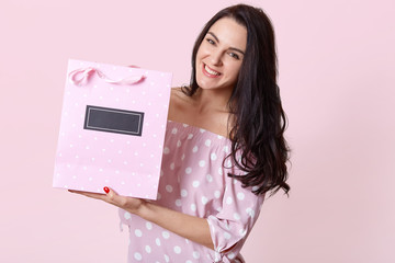 Happy beautiful woman with dark hair, dressed in polka dot dress, holds gift bag with present, has positive facial expression, isolated over light purple background. People and gifts concept