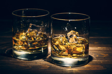 Two glasses of whiskey or another alcohol with ice cubes on wooden background