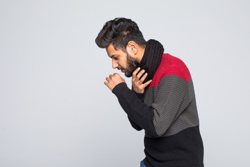Young indian man in gray sweater, scarf sneezing or coughing covering mouth with hands isolated on grey wall background. Healthy lifestyle, ill sick disease treatment cold season concept.