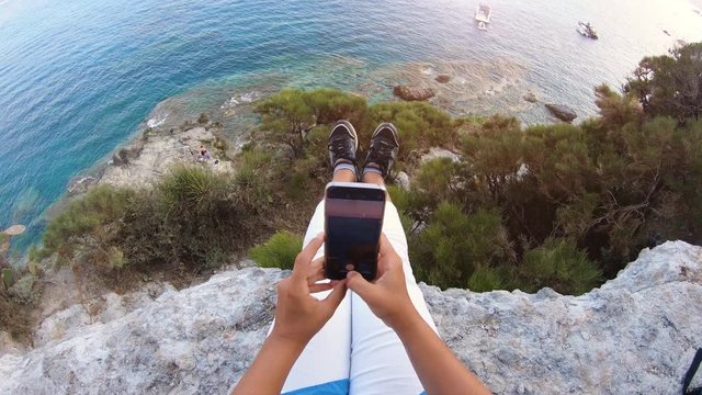 Young woman taking pictures with smartphone sitting on cliff edge looking at the sea from a bay on Ponza Island coast.