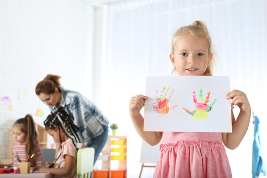 Cute little child showing sheet of paper with colorful hand prints indoors. Painting lesson