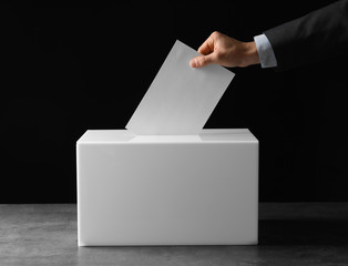Man putting his vote into ballot box on table against black background, closeup
