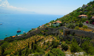 Fortress walls and houses scattered on the green slope descending towards the blue Mediterranean sea with foggy mountains line on the horizon in Alanya city. Antalya, Turkey.