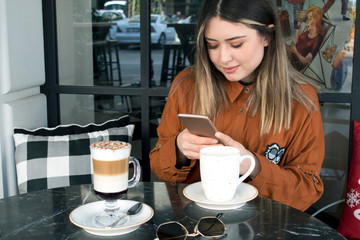 Young woman sitting in coffee shop at table, drinking coffee and using smartphone.