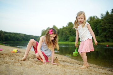 Two young sisters having fun on a sandy lake beach on warm and sunny summer day. Kids playing by the river.