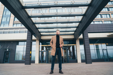 Fototapeta premium Attractive man in trendy brown coat posing near business center. Handsome european man with beard looks away. Concept lifestyle, fashionable men style