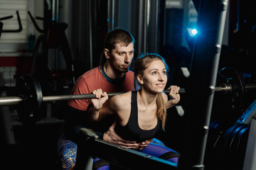 Fototapeta na wymiar Handsome athletic man helps a beautiful girl to squat with a barbell on the shoulders in the gym.