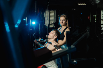Fototapeta na wymiar Active beautiful fitness model girl helps young man to train on the block simulator in the gym.