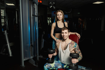 Active beautiful fitness model girl helps to train with dumbbells while sitting to her boyfriend in the gym.