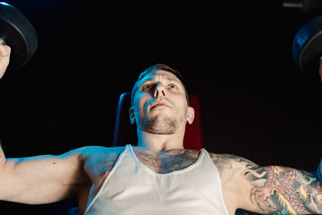 Handsome athletic man trains pectoral muscles by breeding dumbbells while lying in the gym.