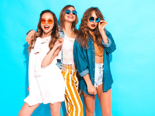 Three young beautiful smiling hipster girls in trendy summer casual dresses. Sexy carefree women posing near blue wall. Positive models going crazy