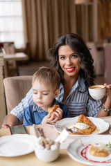 Beautiful young woman with baby son. Light breakfast near window in a cafe. Croissants, omelet, coffee and many different dishes on the table in the cafe. Happy family