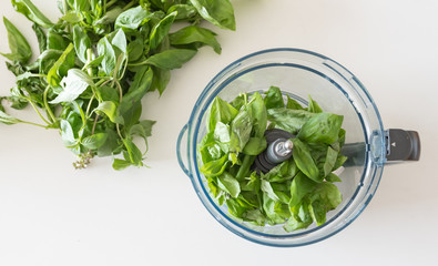 High angle view of basil leaves in food processor bowl with bunch of basil in background on white table (selective focus)