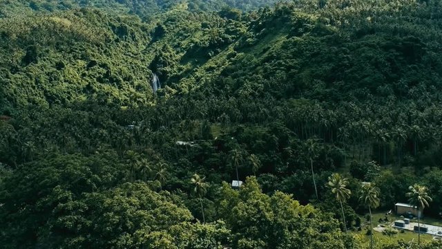 aerial drone footage of a south pacific village on a remote island with sandy beach shore and lush tropical rainforest jungle with waterfall in the background
