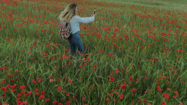 Young pretty woman with blonde hair spins around, makes selfies and photos in middle of beautiful and romantic dreamy poppies field on sunny summer day. Social media and nature concept
