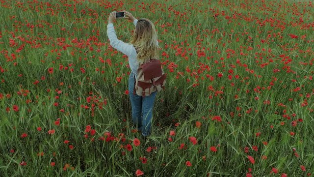 Beautiful teenage millennial girl uses smartphone to make photos of surroundungs, shares memories and experiences of nature around. During hike or field trip in poppy field on picnic