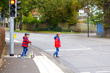 Two little schoolkids boys running and driving on scooter on autumn day. Happy children in colorful clothes and city traffic crossing pedestrian crosswalk with traffic lights.