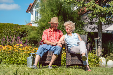 Low-angle view portrait of a happy senior couple in love sitting on chairs, while relaxing together in the garden at home in a sunny day of summer