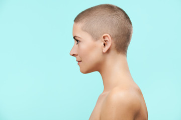 Beautiful mid 30s woman with shaved head studio profile portrait. Model with light nude make-up, isolated on pastel blue background.