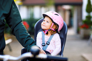 Portrait of little toddler girl with security helmet on the head sitting in bike seat and her father or mother with bicycle. Safe and child protection concept. Family and weekend activity trip.