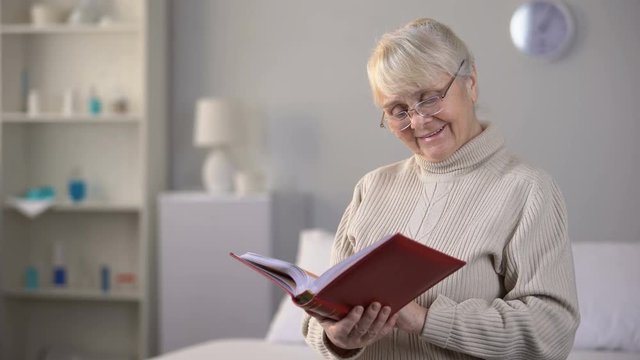 Smiling aged woman in eyeglasses watching photo album, fond memories, family