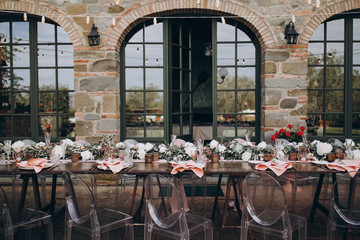 in backyard of villa in Tuscany there is banquet wooden table decorated with cotton and eucalyptus compositions, glasses, candles and plates are placed on table, transparent chairs are next to them