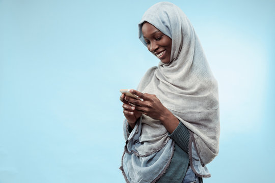 The beautiful young black african muslim girl wearing gray hijab at blue  studio. She standing with mobile phone with a happy smile on her face. The  human emotions, facial expression concept. Trendy Stock Photo | Adobe Stock