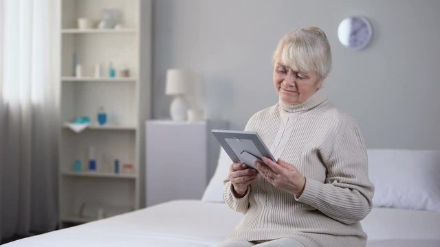 Sad old woman looking at picture of her husband, sitting on bed in living room
