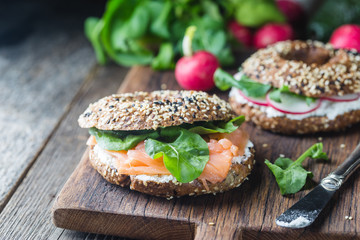 Bagels with cream cheese avocado, fish, arugula and radish on old wooden table. Healthy breakfast food.