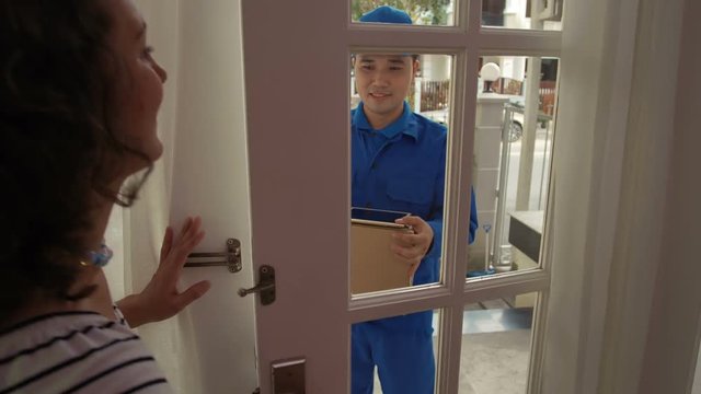 Medium shot of middle-aged woman opening entrance door of her house, taking parcel from Asian deliveryman and signing electronic delivery document with stylus