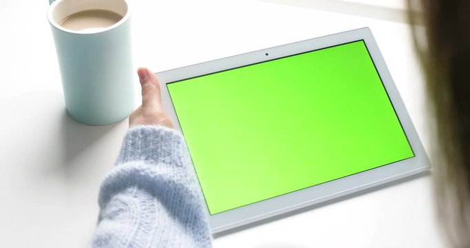 Young girl hold white taplet with green screen on table with coffe or tea cup. Close up 4k footage.