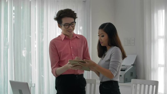 Medium shot of Asian male and female coworkers standing together, looking at tablet screen, discussing funny picture and laughing