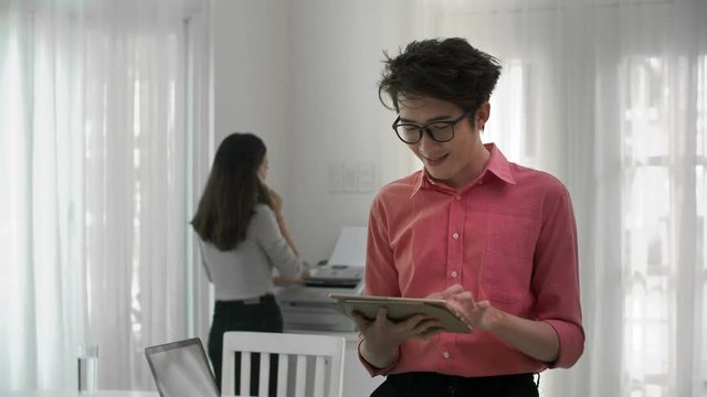 Medium shot of young Asian man standing with tablet in his hands, then looking at camera and smiling, while his female colleague on background printing and having phone talk