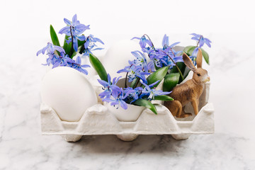 Easter bunny, flowers and eggshell in paper tray on marble background. Spring composition with blue flowers and eggs. Flat lay, top view. Copy Space