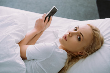 sad woman looking at camera and using smartphone in bed at home