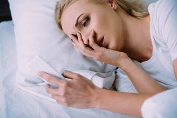 upset woman lying in bed, covering face with hand and using smartphone