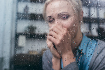 depressed adult woman with folded hands at home through window with raindrops
