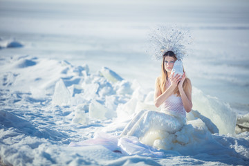 Fototapeta na wymiar Young model in luxurious strapless corset ball gown sitting on slabs of broken ice at the frosty seaside. Winter fairytale concept.