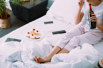 cropped view of woman with cakes and bottle of wine celebrating birthday in bed alone