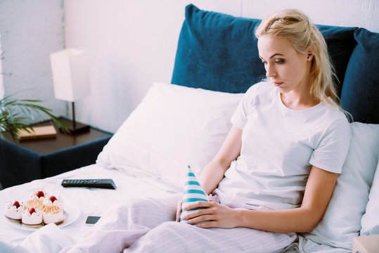 uspet woman holding party hat while celebrating birthday in bed alone