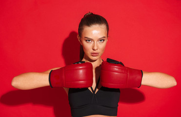 Beautiful young fitness boxer woman posing isolated over red wall background make exercises in gloves.