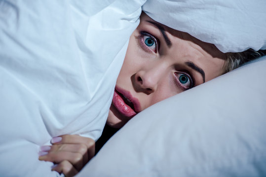 scared woman hiding behind blanket and looking at camera in bedroom at home