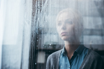 selective focus of sad adult woman at home looking through window with raindrops