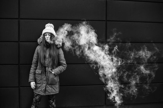 Vape teenager. Young pretty white girl smoking an electronic cigarette opposite modern background on the street in the winter. Bad