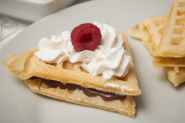 closeup of waffle in shaped heart with chantilly cream and raspberry in a white plate